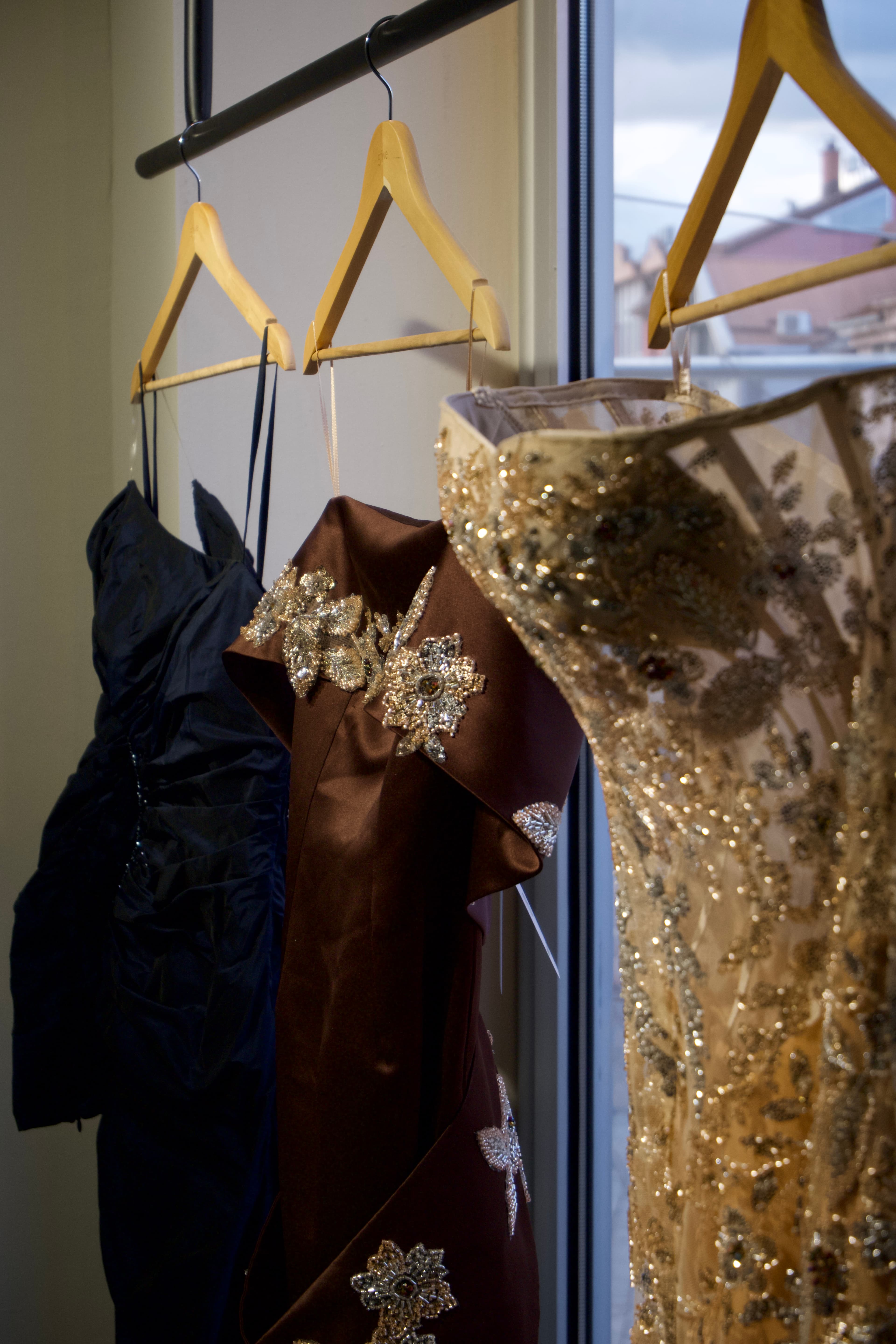 Three RABEL gowns hanging near the boutique window — navy, rust, and gold crystal — at the showroom opening